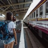Tânără cu un rucsac care așteaptă trenul pe peronul unei gări, privind în depărtare