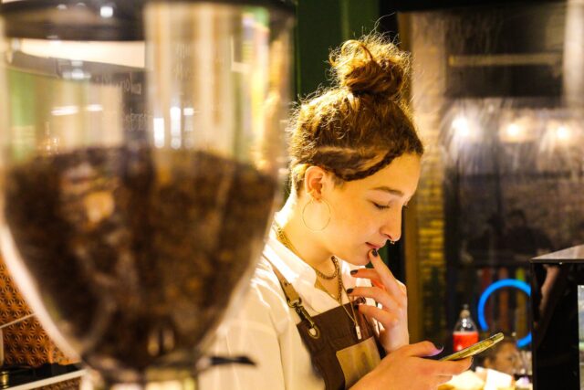 Student care lucrează part-time ca barista într-o cafenea, pregătind o cafea pentru un client